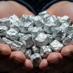 Mãos segurando vários blocos metálicos prateados e irregulares, representando o elemento Califórnio-252 em estado bruto.