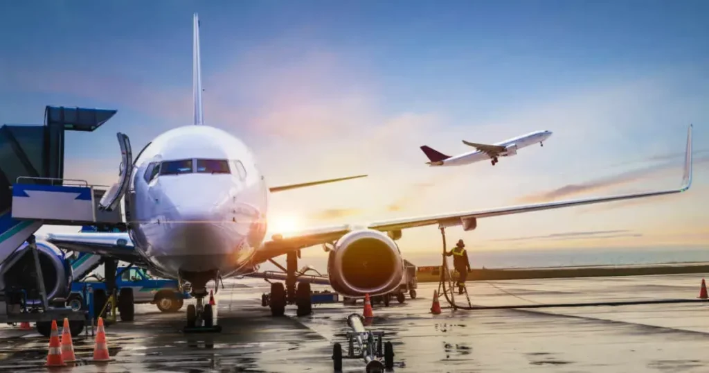 Avião estacionado sendo abastecido em aeroporto ao pôr do sol, com outro avião decolando ao fundo. Cena simboliza segurança e tecnologia na aviação.