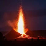Erupção vulcânica intensa durante a noite, com jato de lava incandescente sendo expelido do topo de um vulcão e escorrendo pelas encostas. Pessoas observam de longe em silhueta. Tesouro Escondido