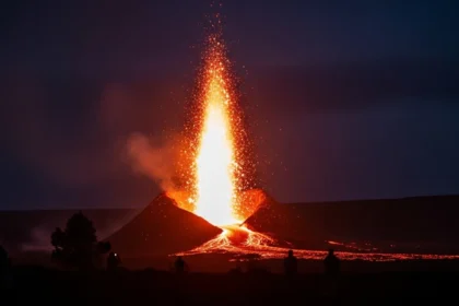 Erupção vulcânica intensa durante a noite, com jato de lava incandescente sendo expelido do topo de um vulcão e escorrendo pelas encostas. Pessoas observam de longe em silhueta. Tesouro Escondido