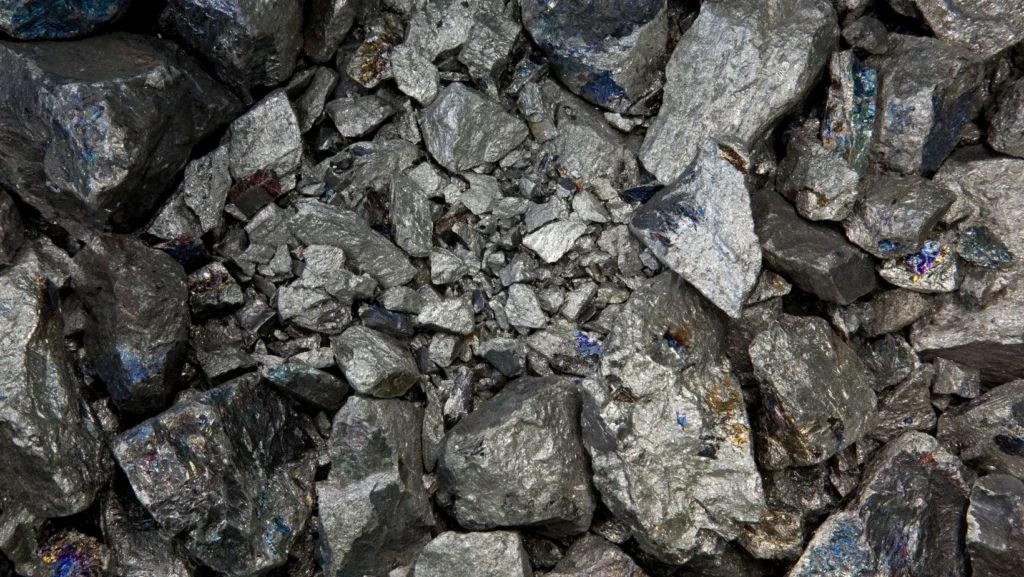 Foto bem de perto de um monte de nióbio bruto, que é um minério. As pedras são cinzas e prateadas, com um brilho de metal forte, e umas partes coloridas que parecem um arco-íris.
