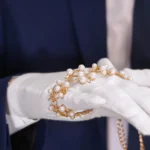 Mãos de um joalheiro usando luvas brancas, segurando e avaliando cuidadosamente um bracelete de ouro com pérolas.