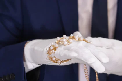 Mãos de um joalheiro usando luvas brancas, segurando e avaliando cuidadosamente um bracelete de ouro com pérolas.