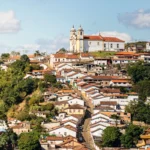 Vista panorâmica da cidade histórica de Ouro Preto, Minas Gerais. A imagem mostra um morro coberto por casarões coloniais com telhados de terracota e paredes predominantemente brancas. No topo do morro, uma igreja barroca branca com detalhes em ocre e duas torres se destaca contra um céu azul com nuvens brancas. A vegetação verde e densa cerca as construções. Rota do Ouro.