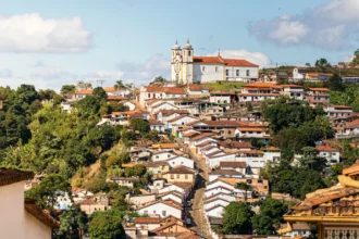 Vista panorâmica da cidade histórica de Ouro Preto, Minas Gerais. A imagem mostra um morro coberto por casarões coloniais com telhados de terracota e paredes predominantemente brancas. No topo do morro, uma igreja barroca branca com detalhes em ocre e duas torres se destaca contra um céu azul com nuvens brancas. A vegetação verde e densa cerca as construções. Rota do Ouro.