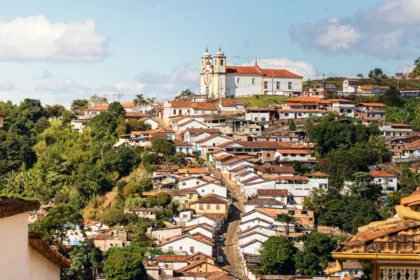 Vista panorâmica da cidade histórica de Ouro Preto, Minas Gerais. A imagem mostra um morro coberto por casarões coloniais com telhados de terracota e paredes predominantemente brancas. No topo do morro, uma igreja barroca branca com detalhes em ocre e duas torres se destaca contra um céu azul com nuvens brancas. A vegetação verde e densa cerca as construções. Rota do Ouro.