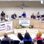 Audiência pública em Minas Gerais sobre a Ferrovia da Cedro, com moradores de Mateus Leme e região presentes, segurando cartazes de protesto contra o projeto.